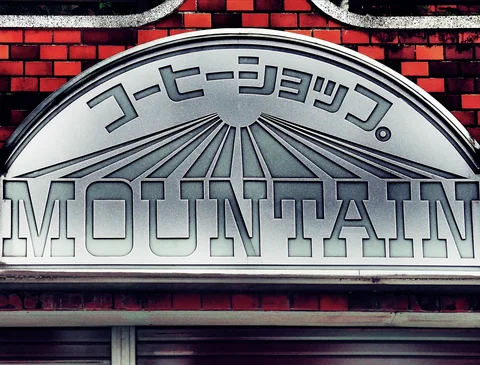 An arched silver MOUNTAIN coffee shop sign with radiating lines sits against red brickwork.