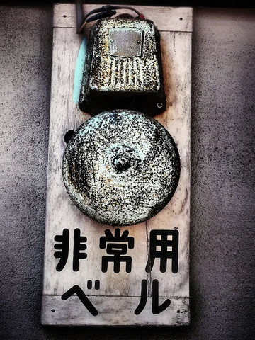 A corroded round alarm bell and small rectangular fixture sit above dark lettering on a pale board.