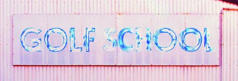 Faded blue lettering spells Golf School across a pale rectangular sign on pink corrugated siding.