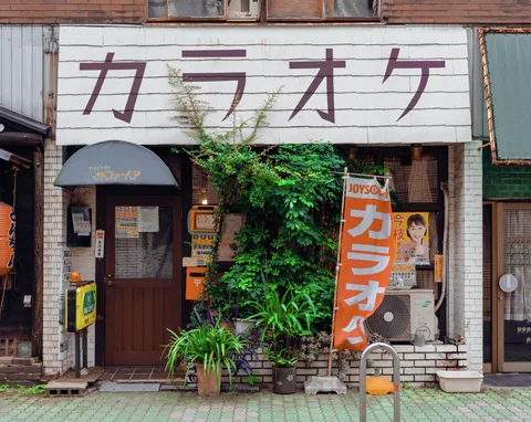 Large Japanese lettering spans a small storefront with an orange banner and dense plants beside its entrance.