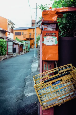 An orange mailbox hangs above leaning yellow plastic racks beside a quiet narrow residential lane.