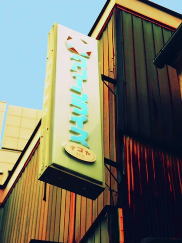 A tall pale projecting sign with raised turquoise Japanese lettering hangs beside dark corrugated siding.