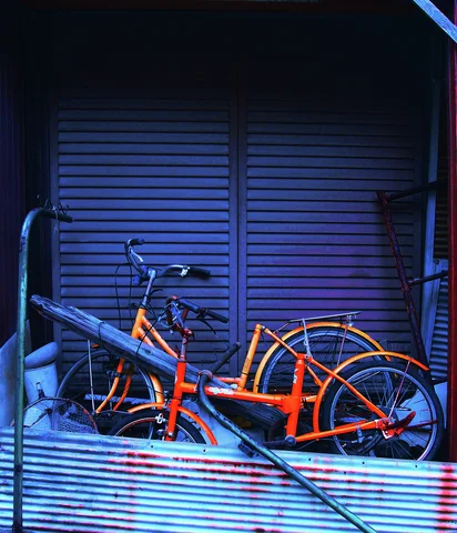 An orange bicycle rests behind weathered corrugated panels before closed blue shutters.