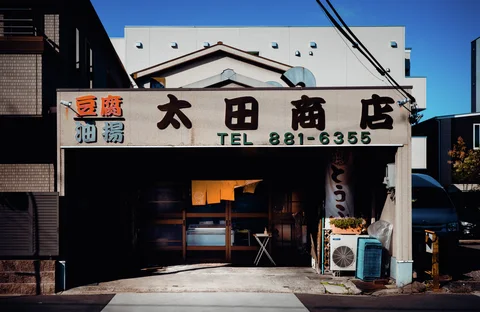 A small open-front shop has a wide sign, yellow curtains, and an outdoor air conditioner.