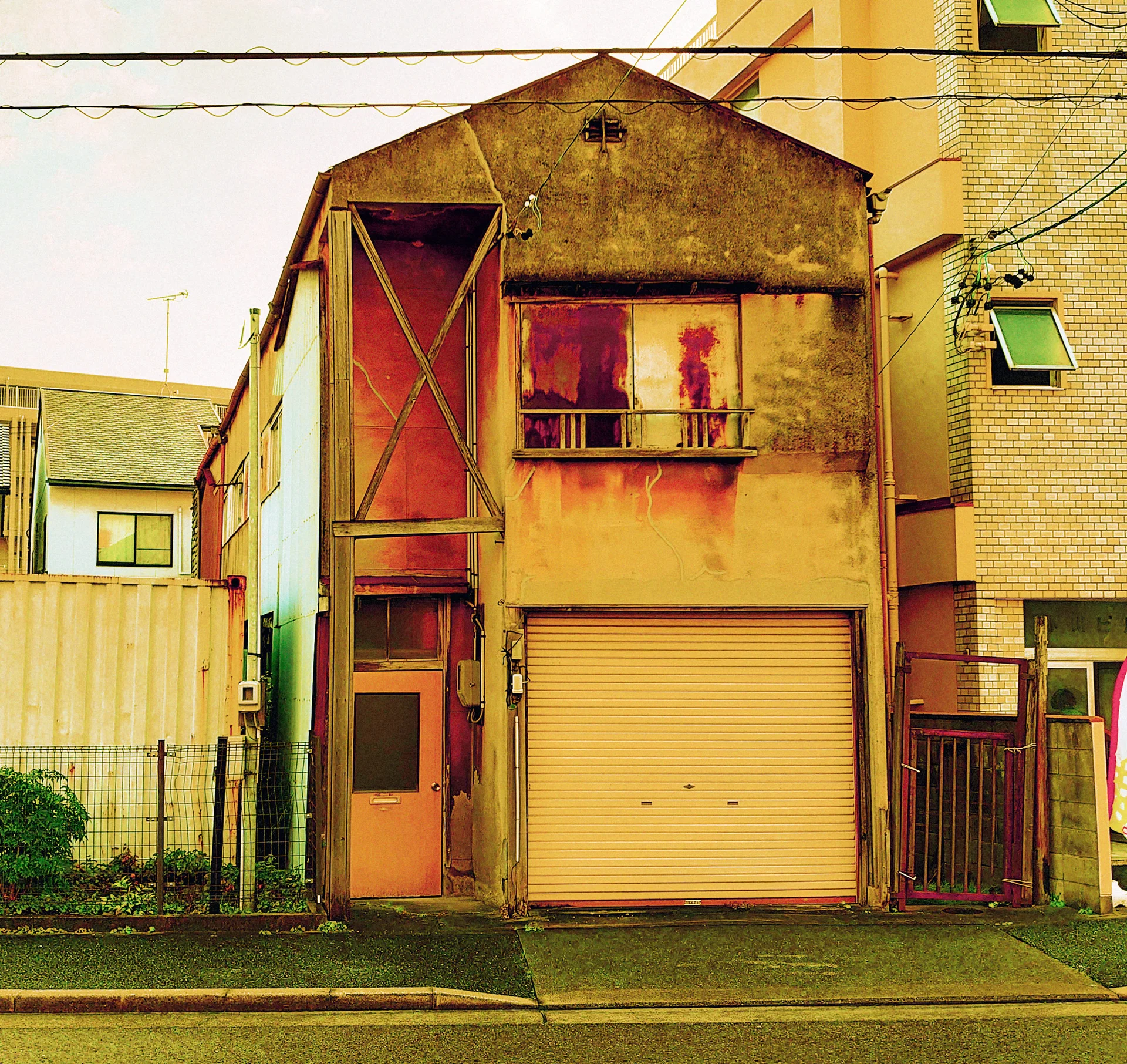 A narrow weathered yellow building has a peaked facade, boarded upper window, and closed ground-floor shutter.