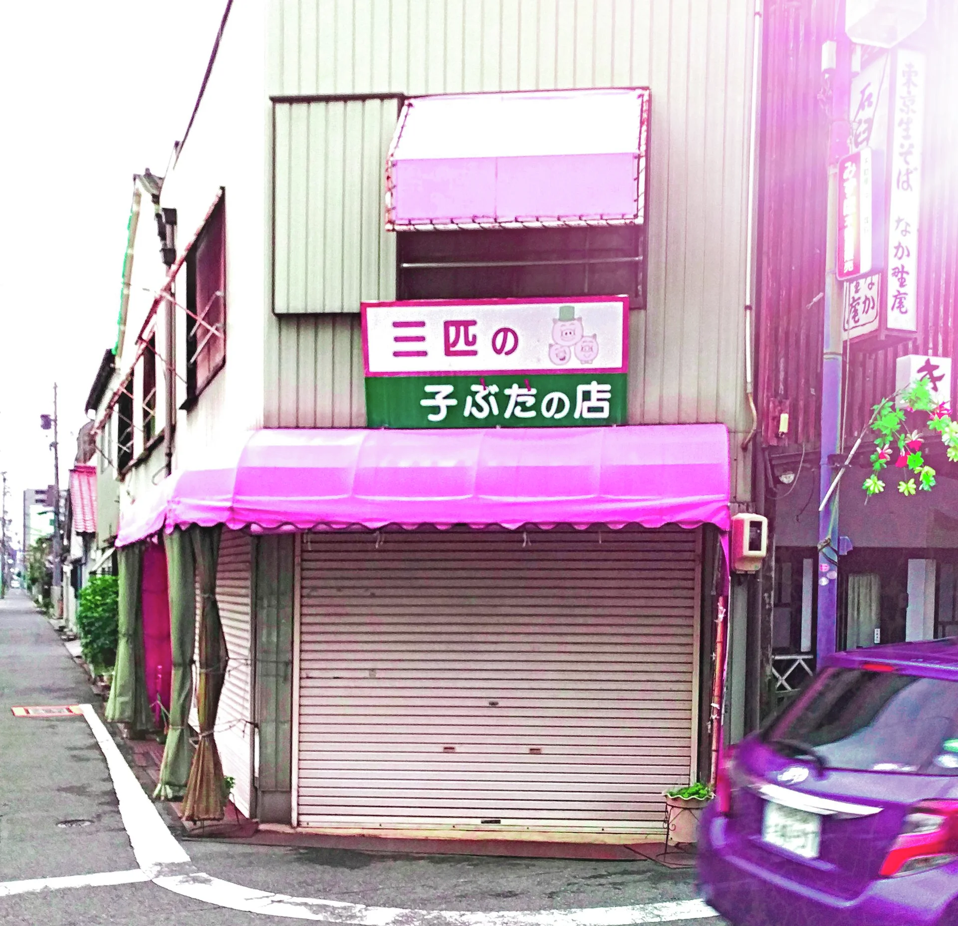 A pink awning shades closed corner-shop shutters beside a purple car and narrow lane.