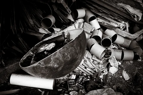 Short pipes and broken debris surround a dented metal bowl in a scrap pile in black and white.