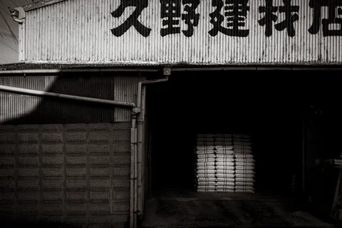 Bold Japanese lettering spans a corrugated warehouse facade above a shadowy open bay in black and white.