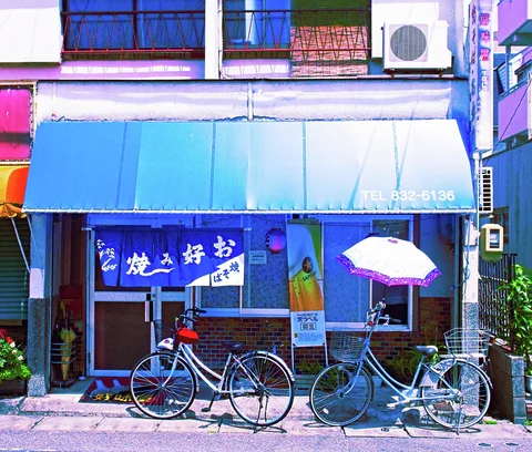 Two bicycles stand outside a blue-fronted shop beneath an awning and a small pale umbrella.