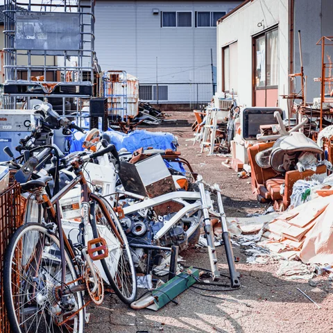 Bicycles, vehicle parts and assorted scrap crowd a sunlit yard between industrial buildings.
