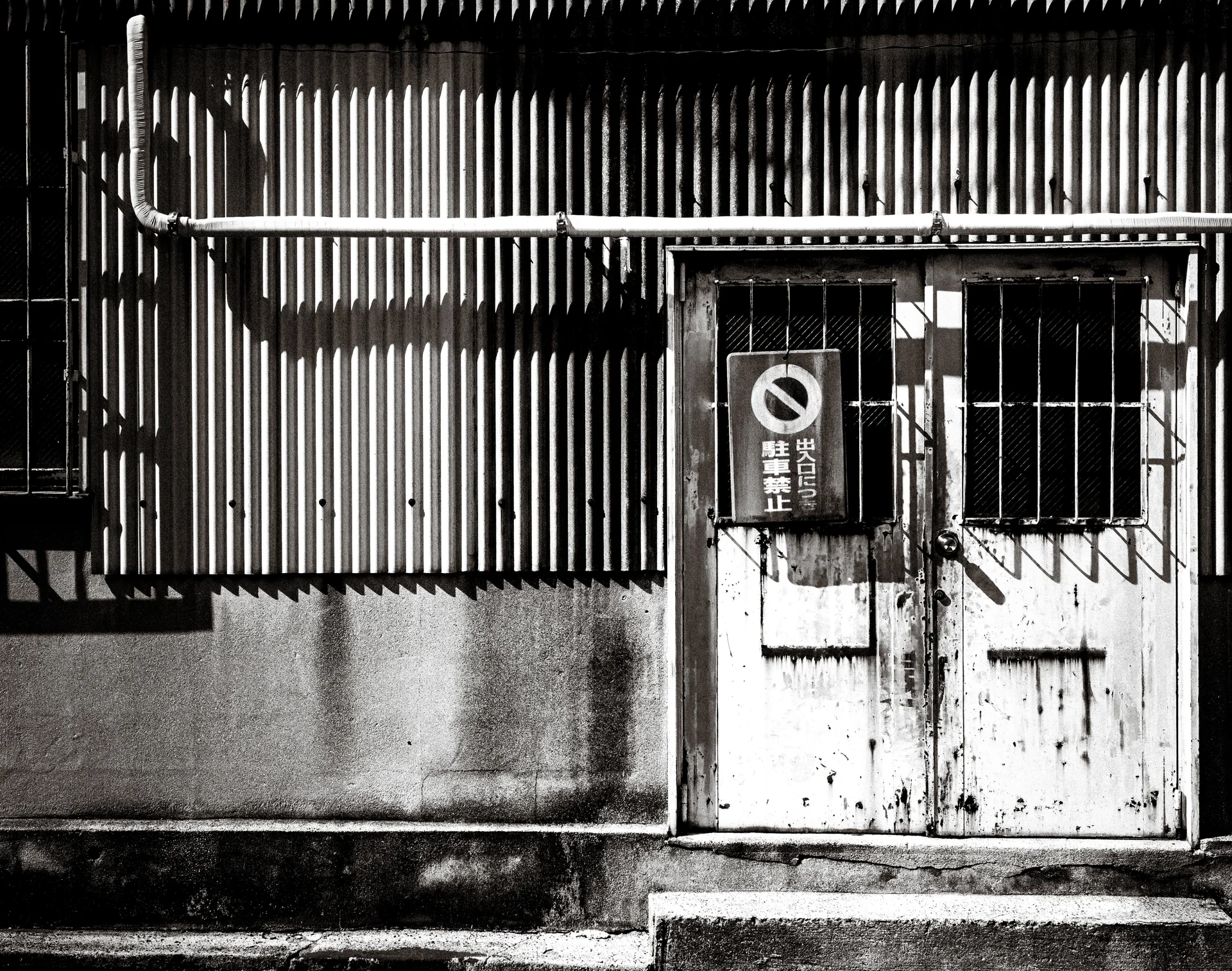 Weathered white double doors with barred windows stand in a corrugated metal wall in monochrome.