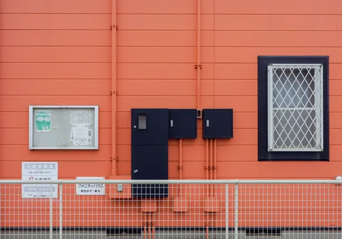 Dark utility boxes and a barred window contrast with orange horizontal siding behind a low wire fence.