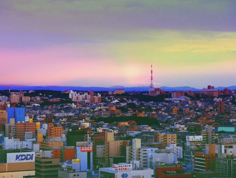 A slender red-and-white tower rises over colorful city buildings beneath a pastel pink and yellow sky.