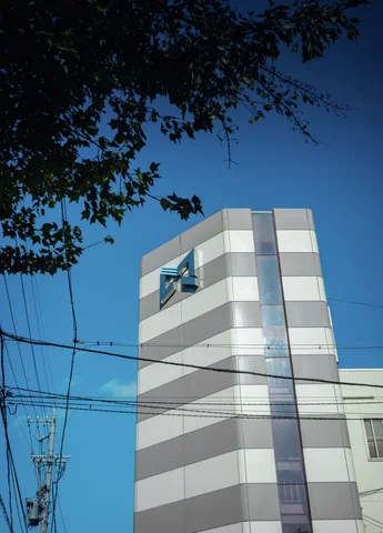 A narrow striped tower rises beneath dark leafy branches and crossing wires against a deep blue sky.