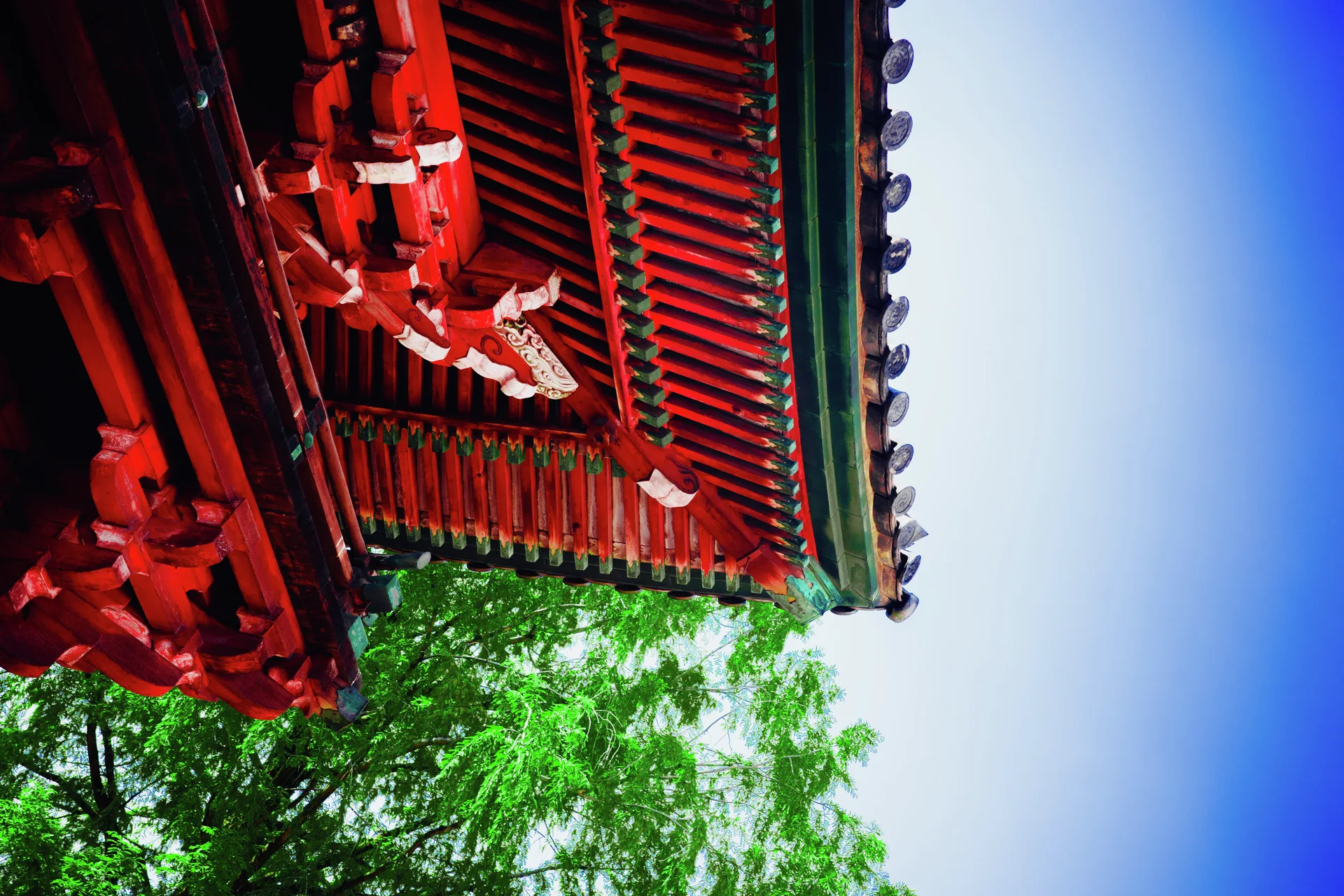 Red and green painted roof brackets project above leafy branches beneath a bright blue sky.