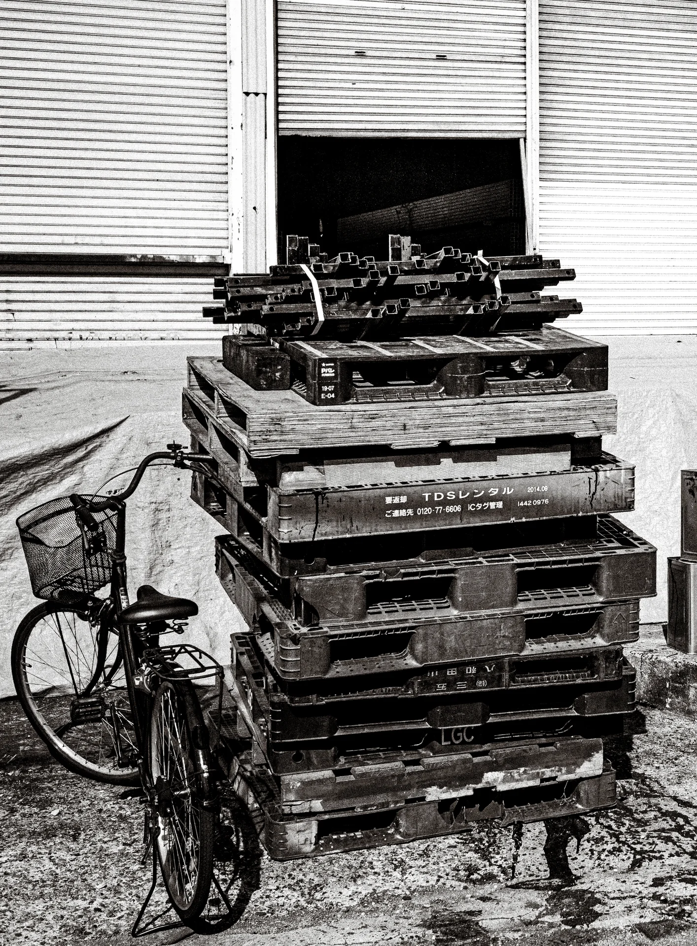A bicycle leans beside stacked wooden pallets below closed metal shutters in black and white.