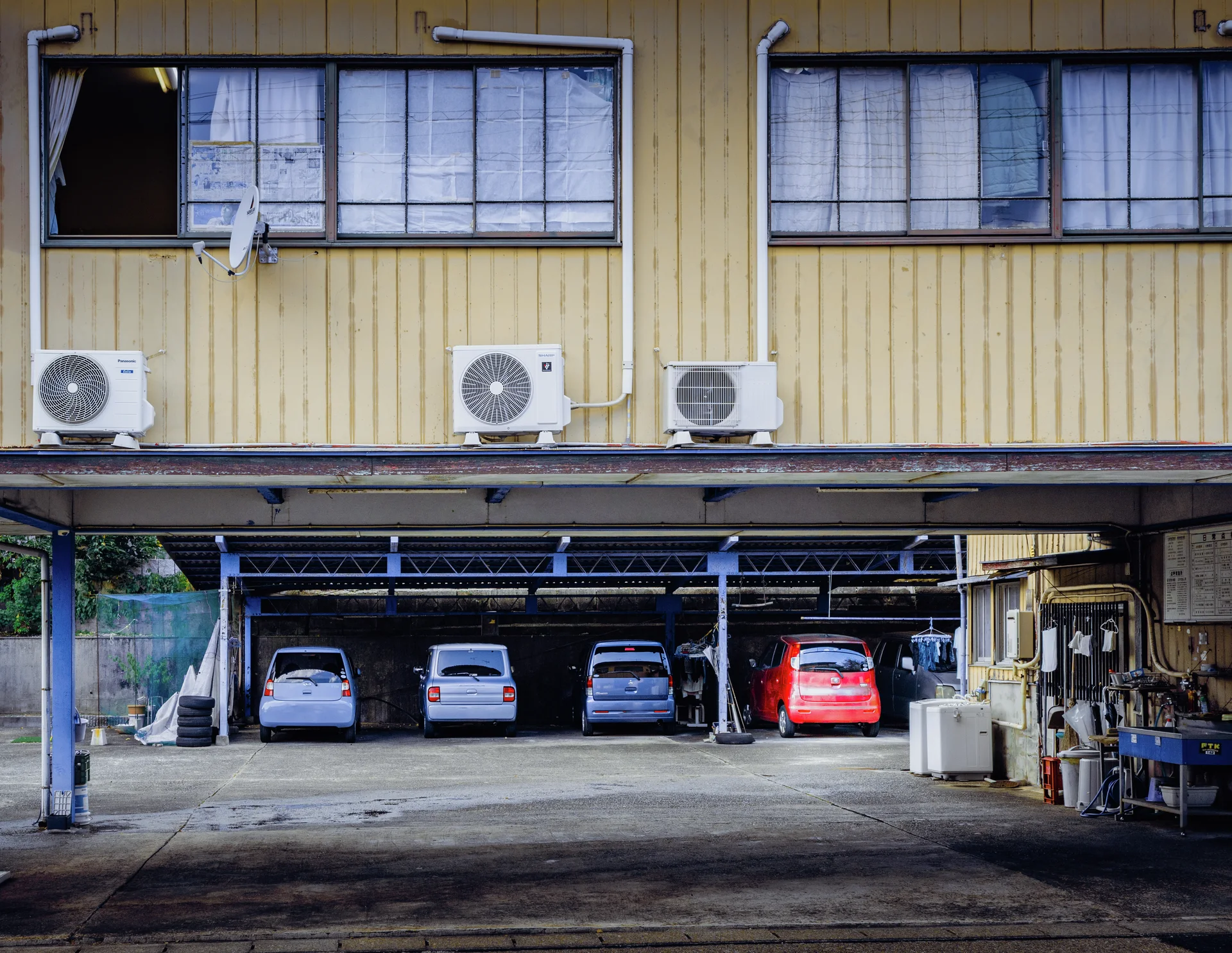 Four cars are parked beneath a yellow upper floor lined with windows and outdoor air conditioners.