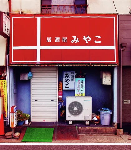A large red sign hangs above a closed shutter and air-conditioning unit outside a small storefront.