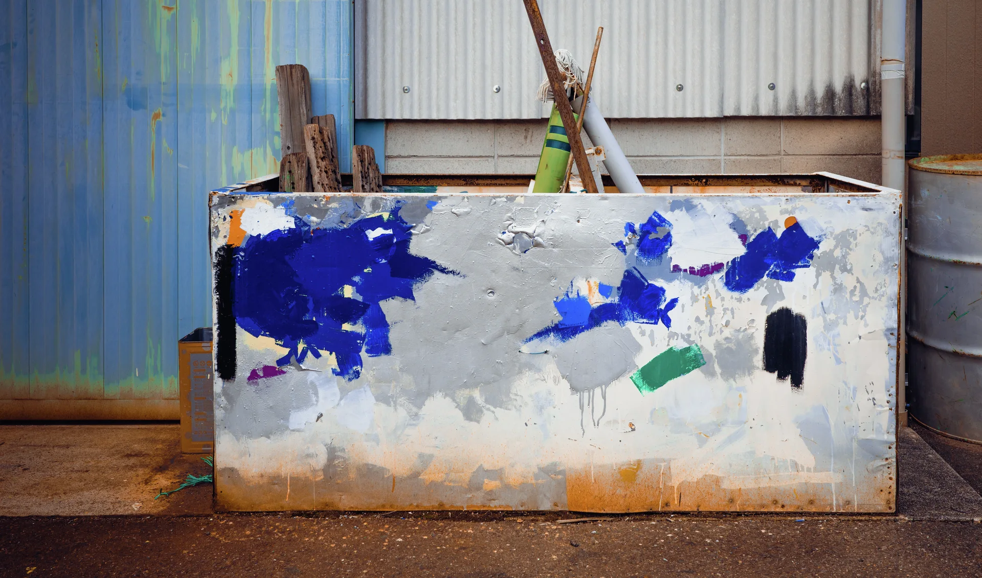 A peeling painted metal panel leans against corrugated walls beside upright pieces of wood.