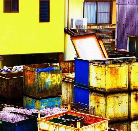 Stacks of yellow and blue metal bins crowd a yard beside a bright yellow building.