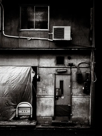 Exposed pipes cross a weathered wall above a narrow door beside a tarp-covered object.