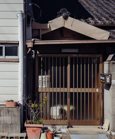 A wooden slatted entrance sits beneath a small tiled canopy beside potted plants and a mailbox.