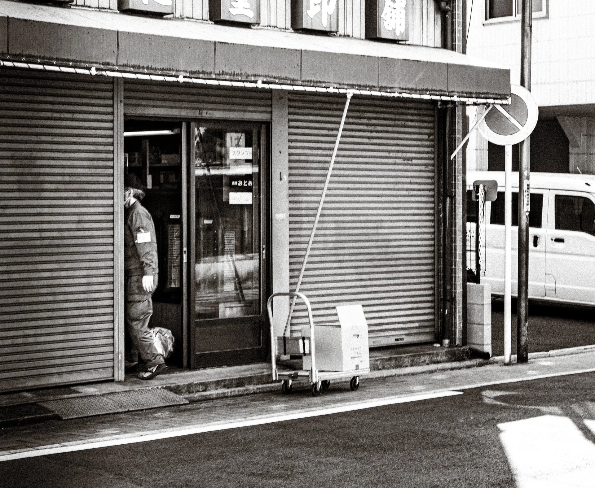 A person stands in a shop doorway between closed shutters beside a parked van in black and white.