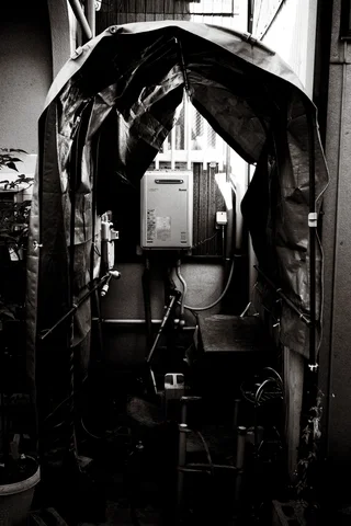 A dark tarp arches over equipment in a tight gap between buildings in black and white.