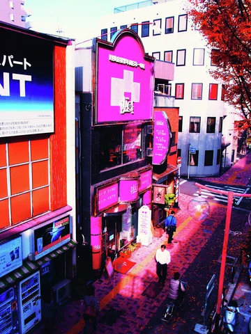 Pedestrians move along a narrow brick-paved shopping street beneath vivid pink signs and colorful facades.