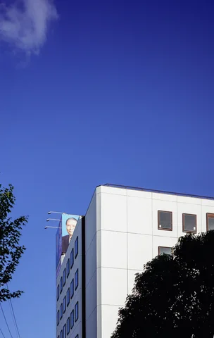 A pale building corner rises above dark tree foliage beneath an expansive deep blue sky.