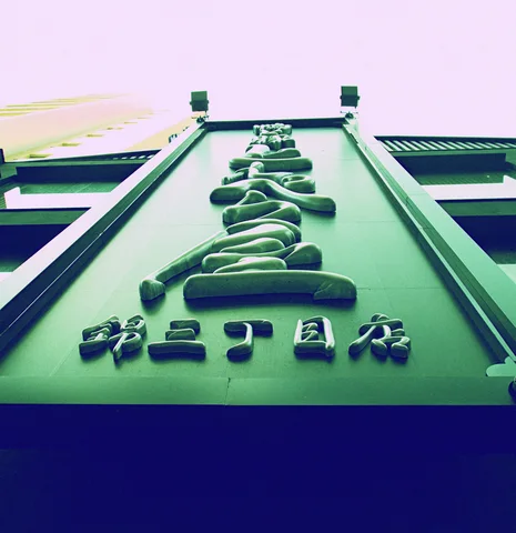Raised dark characters climb a tapered green facade beneath two small rooftop posts.