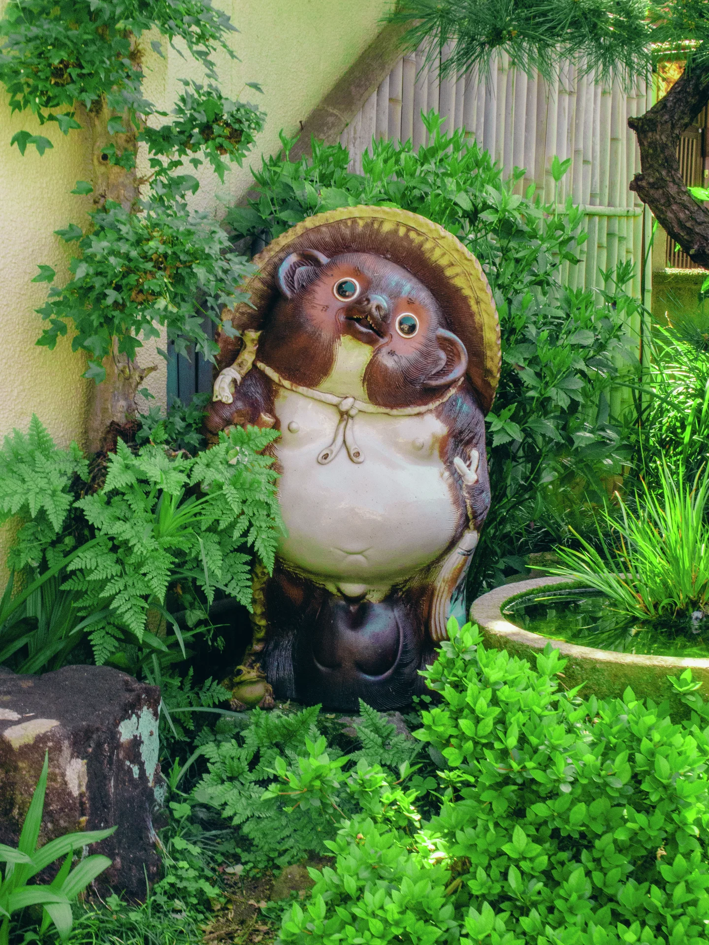 A round-bellied animal statue wearing a straw-colored hat stands surrounded by lush green plants.