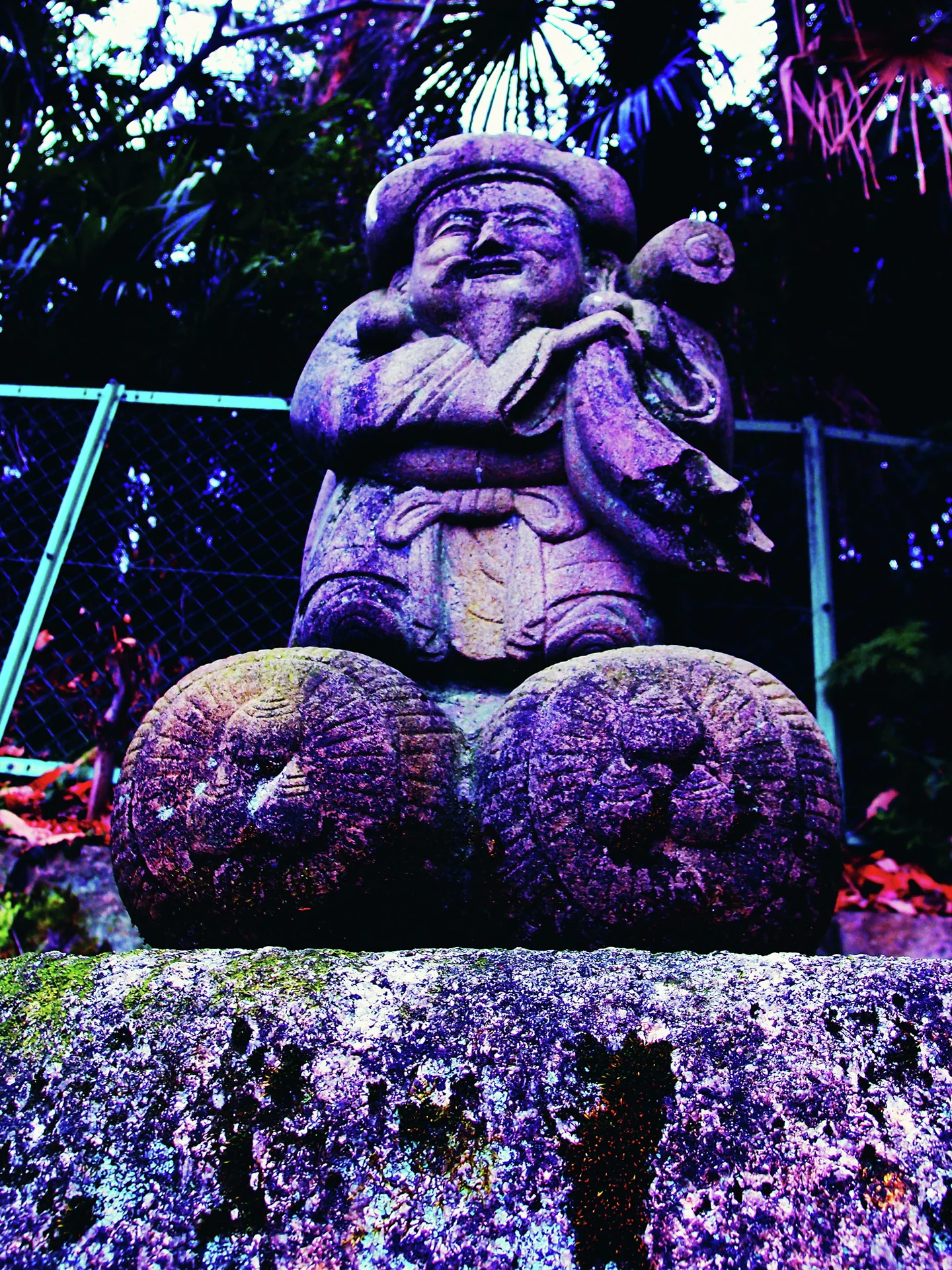 A smiling stone figure holds a sack and mallet above two rounded bales beneath dark foliage.