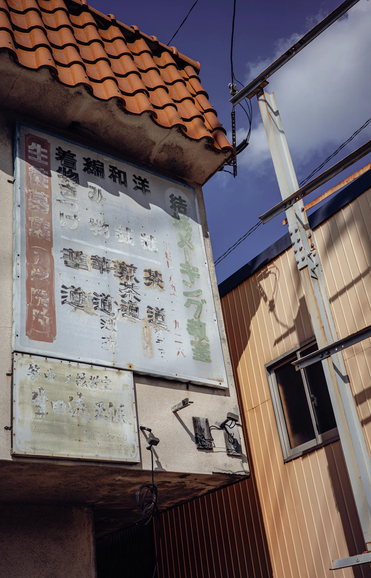 A weathered vertical sign sits beneath orange roof tiles beside overhead cables and corrugated buildings.