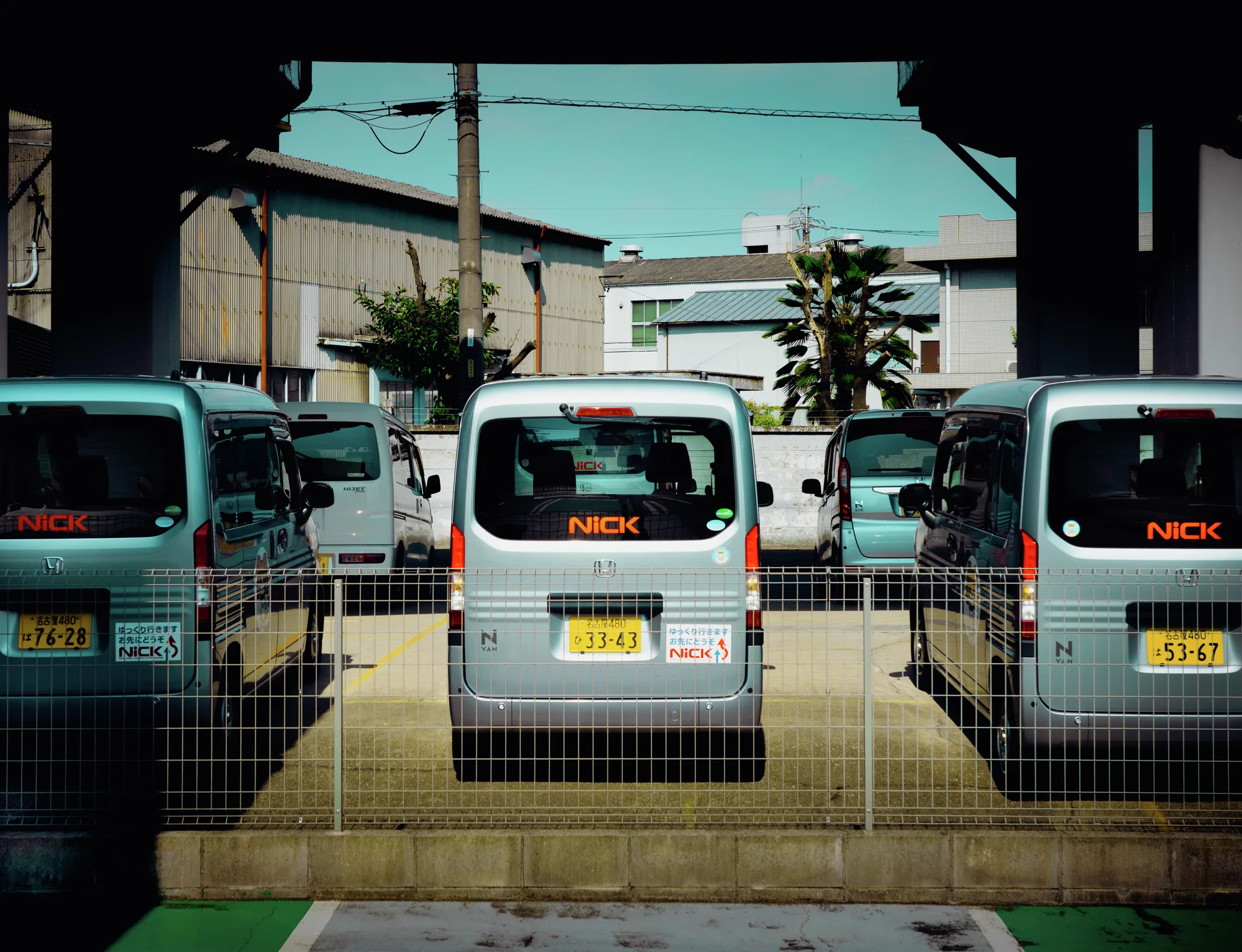 Several pale vans with orange rear lettering sit behind a wire fence in a parking area.