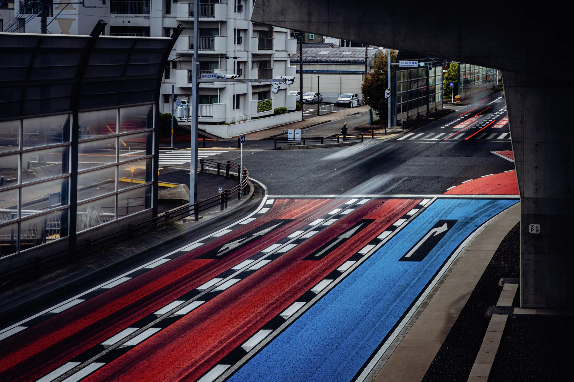 Red and blue road markings curve through an intersection beneath an elevated roadway and transparent barriers.