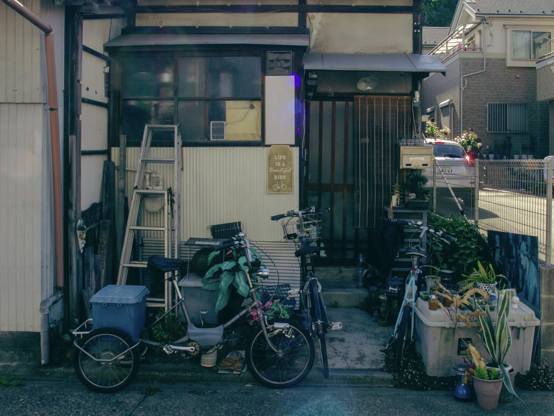Bicycles, a leaning ladder and assorted containers crowd the small entrance of a weathered house.