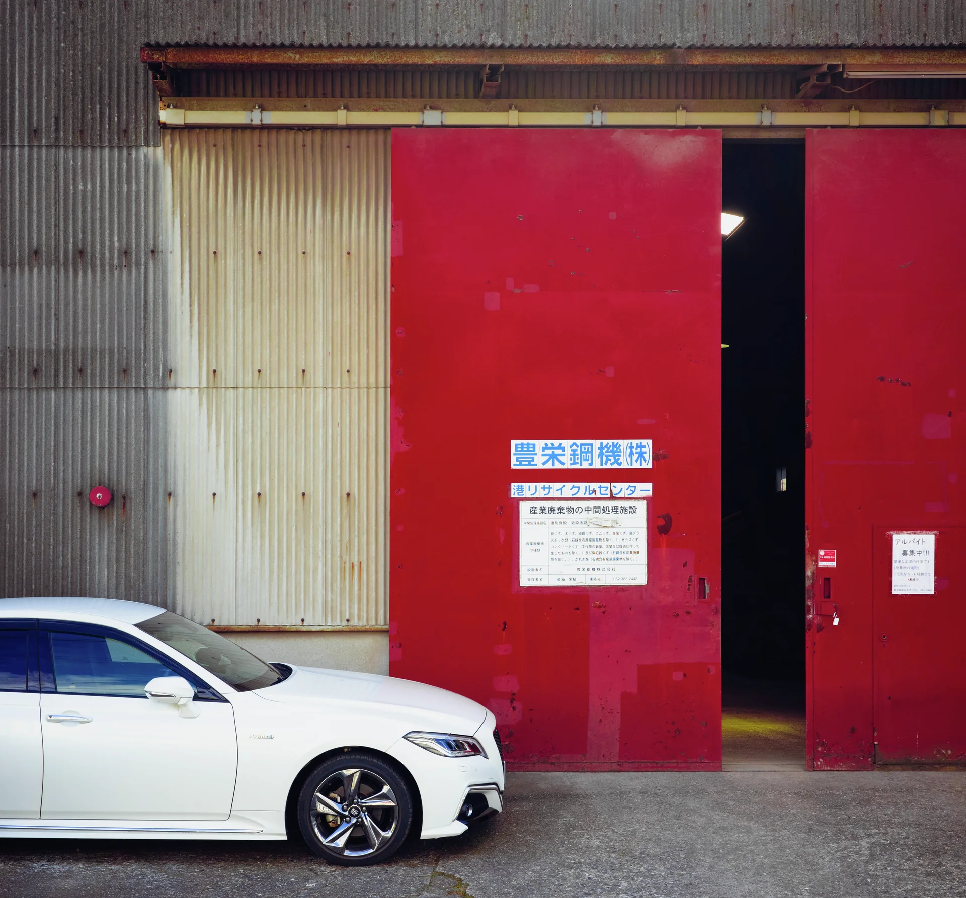 A white sedan sits beside tall red sliding doors with a narrow dark opening.
