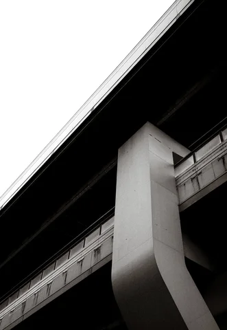 A tall curved concrete support meets a dark elevated roadway in a sharply angled black and white view.