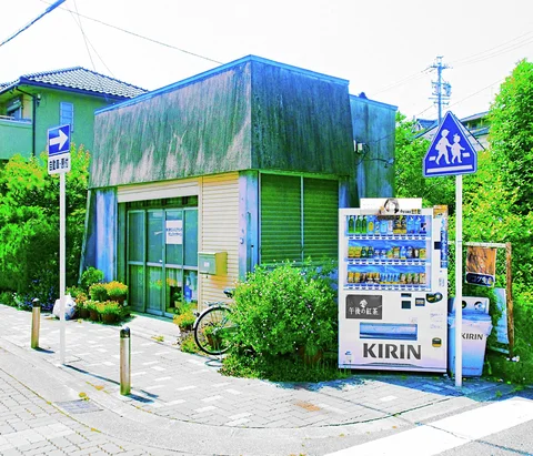 A drinks vending machine stands beside a low weathered blue-green shop at a sunny street corner.