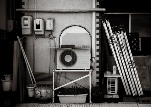 Air conditioners and utility boxes share a wall with leaning ladders and planters in black and white.