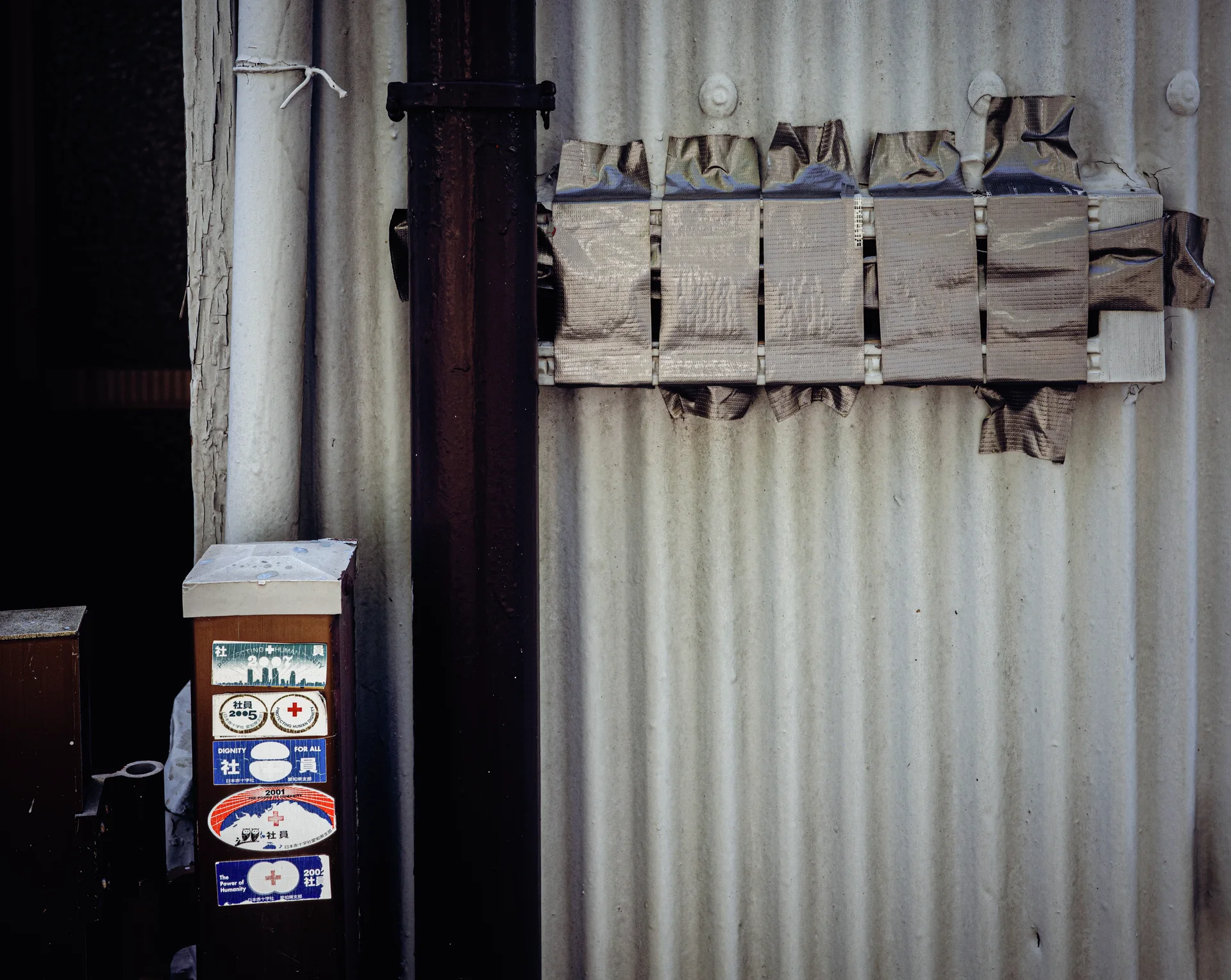 A small window is covered with rough boards and fabric on a pale corrugated wall beside a stickered box.