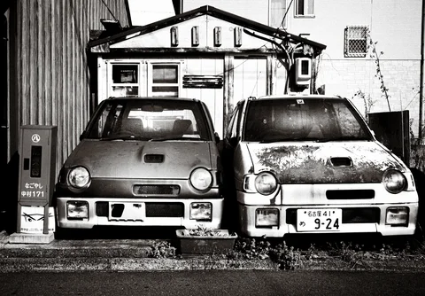 Two small weathered cars stand tightly side by side before a modest building in black and white.