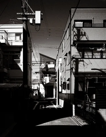 Sunlight illuminates a narrow residential alley between dark buildings and projecting balconies in black and white.