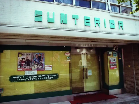 Raised turquoise lettering spans a pale shop facade above broad yellow-backed windows covered with notices.