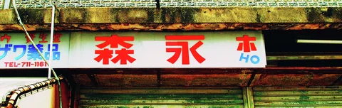 Large red Japanese characters span a weathered white sign above closed shutters beneath a stained concrete ledge.
