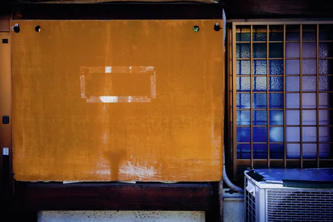 An orange panel with a pale rectangular mark stands beside a blue barred window and air conditioner.