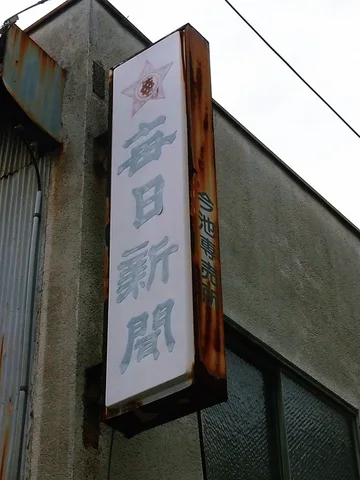 A narrow weathered vertical sign with faded blue characters projects from a gray building beneath overhead wires.