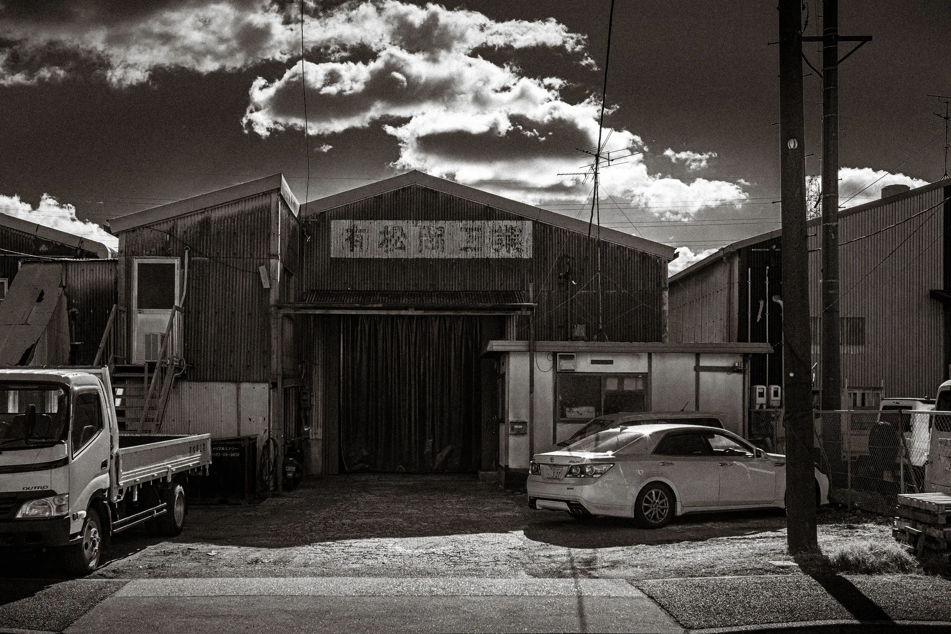 A truck and white sedan stand outside a weathered industrial building beneath dramatic monochrome clouds.