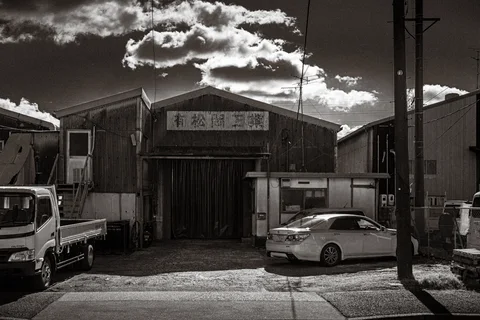 A truck and white sedan stand outside a weathered industrial building beneath dramatic monochrome clouds.